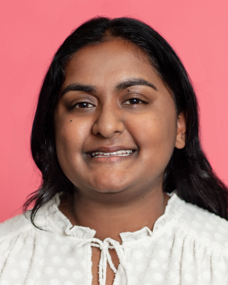 A woman with long dark hair and a white blouse smiles in front of a pink background.